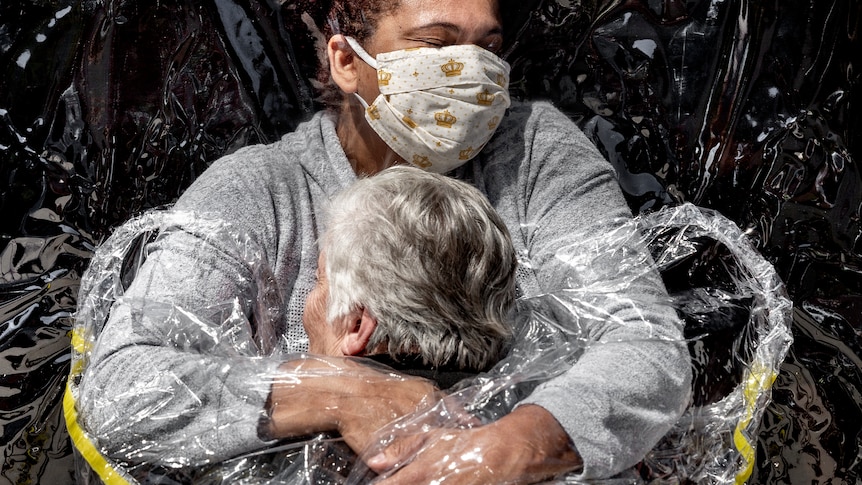 An old woman is embraced by a nurse through a plastic sheet.