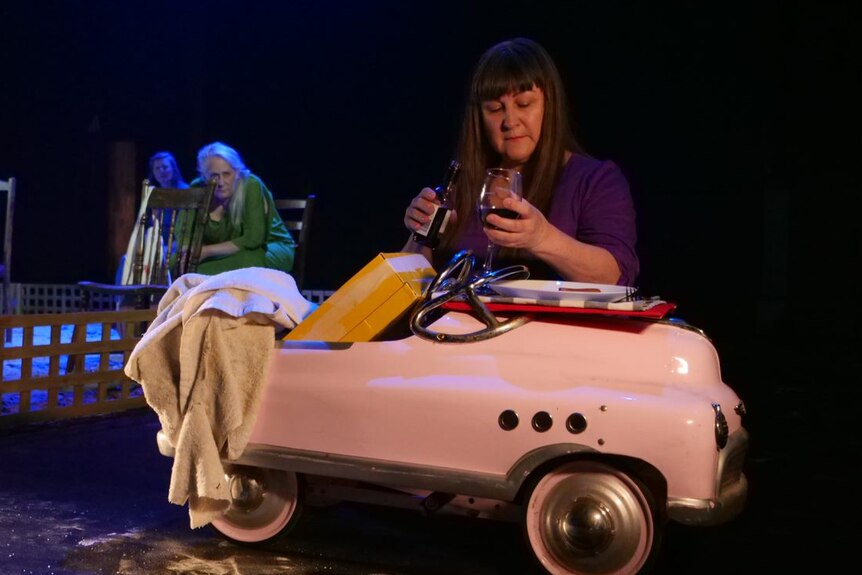 A woman pours a glass of wine in front of a prop car on stage.