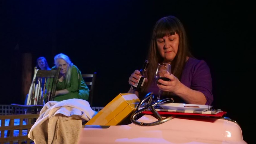 A woman pours a glass of wine in front of a prop car on stage.