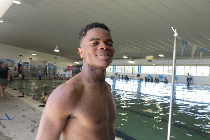 A young muscular African man is bare chested as he stands next to an indoor swimming pool.
