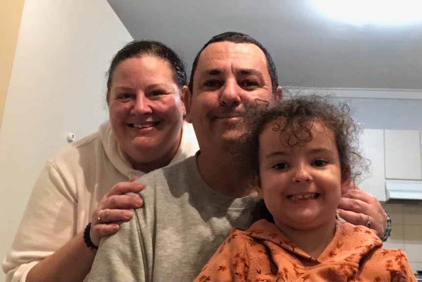 A picture of a couple with their daughter smiling for a close-up selfie indoors.