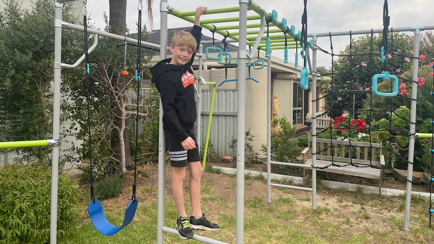 boy posing in front of monkey bars, arm outstretched holding bar above his head.
