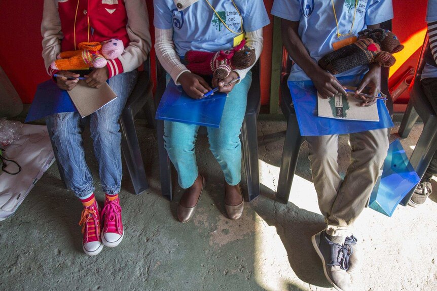 Three girls, photographed from the shoulders down, hold knitted dolls as they sit on plastic chairs.