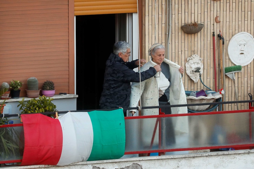 An elderly man helps an elderly woman put on an overcoat on a balcony with Italian flag behind them.