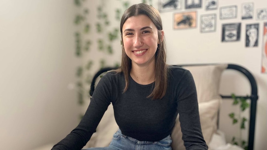 A teenage girl sits cross legged on a bed wearing a black long sleeved shirt and blue jeans with a book open in front of her.