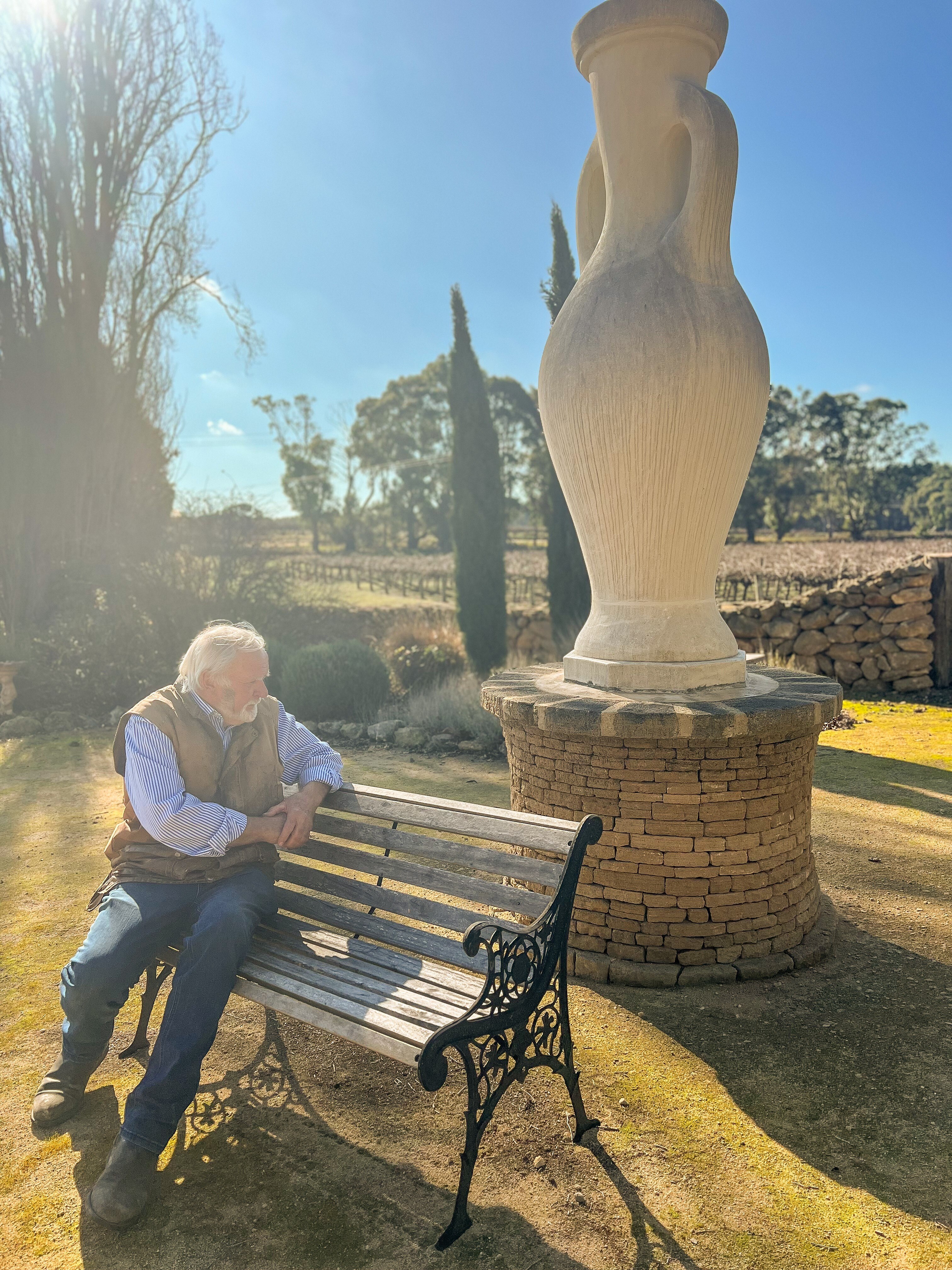 A man with grey hair sits on a bench looking back at a drystone sculpture