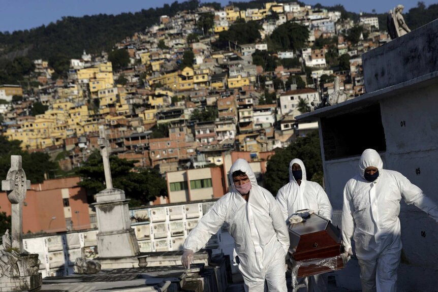 Gravediggers dressed in a white full bodysuit carry a coffin against a backdrop of houses on a hill.