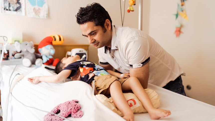 Qasim Butt looks over his son who is lying on a bed with medical equipment around him.