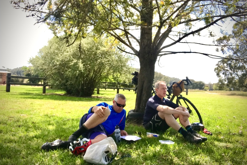 Two men in lycra sit under a tree and rest.
