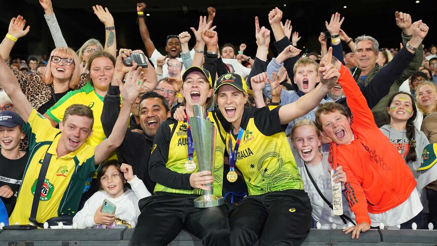 Two Australian women's cricketers hold the T20 World Cup trophy as fans go wild behind them.