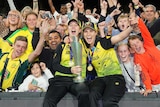 Two Australian women's cricketers hold the T20 World Cup trophy as fans go wild behind them.