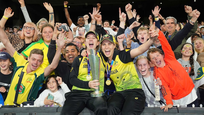 Two Australian women's cricketers hold the T20 World Cup trophy as fans go wild behind them.