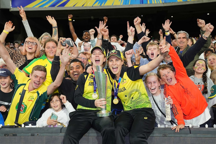 Two Australian female cricketers hold up the T20 World Cup trophy as fans go wild behind them.