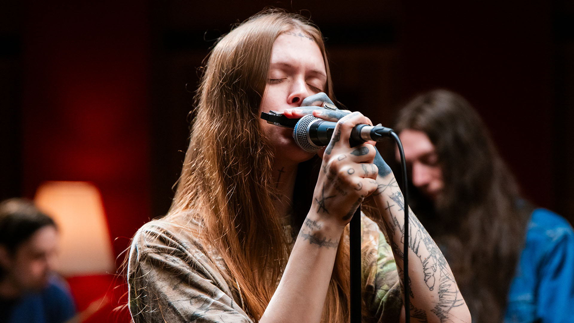 A woman with long, straight brown hair and heavy tattoos plays the harmonica into a microphone with her eyes closed