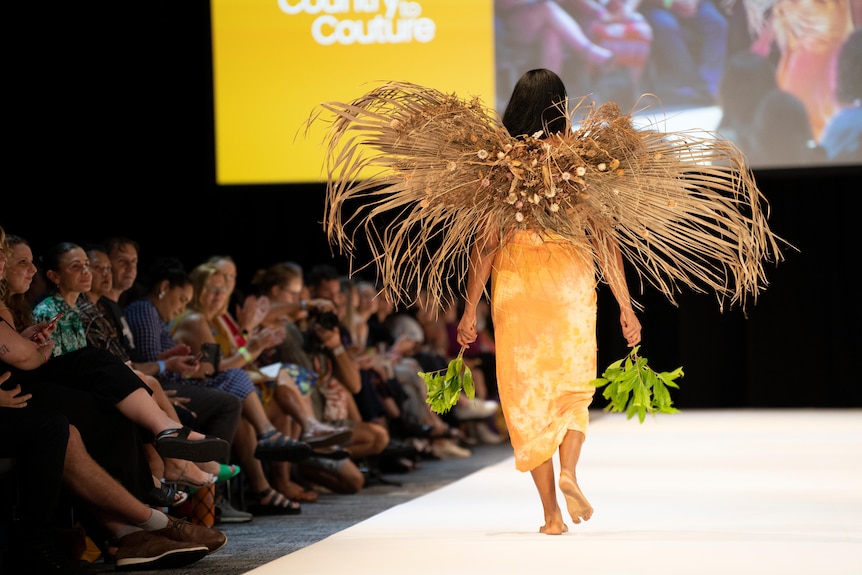 A woman walks away from the camera on a fashion runway. She wears wings made out of grass.