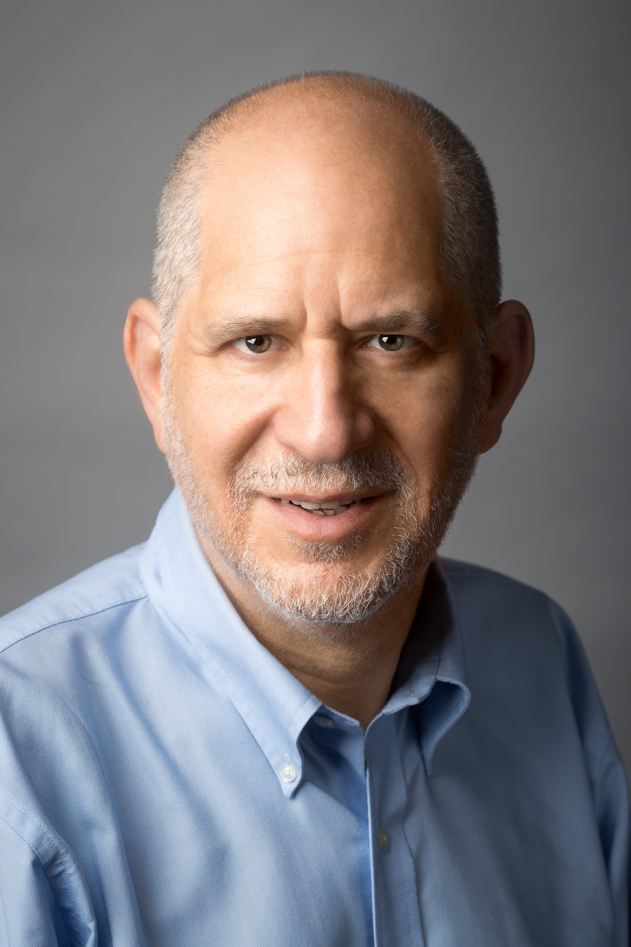 Portrait of an older man with grey stubble and a light blue button up shirt.