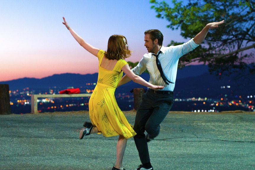 A film still of Stone and Gosling dancing together at sunset. Stone is in a yellow dress, Gosling in a dress shirt and tie
