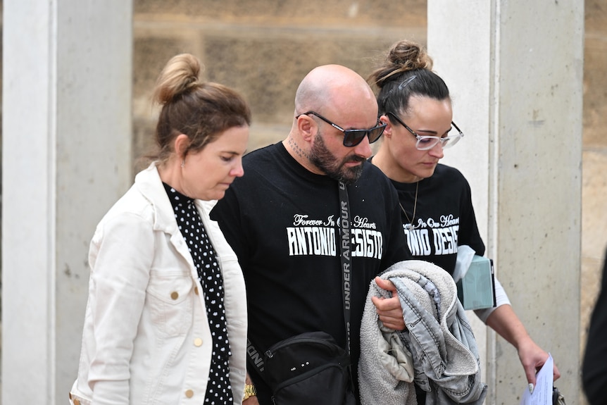 A man wearing sunglasses and a black t-shirt walks alongside two women outside a court.