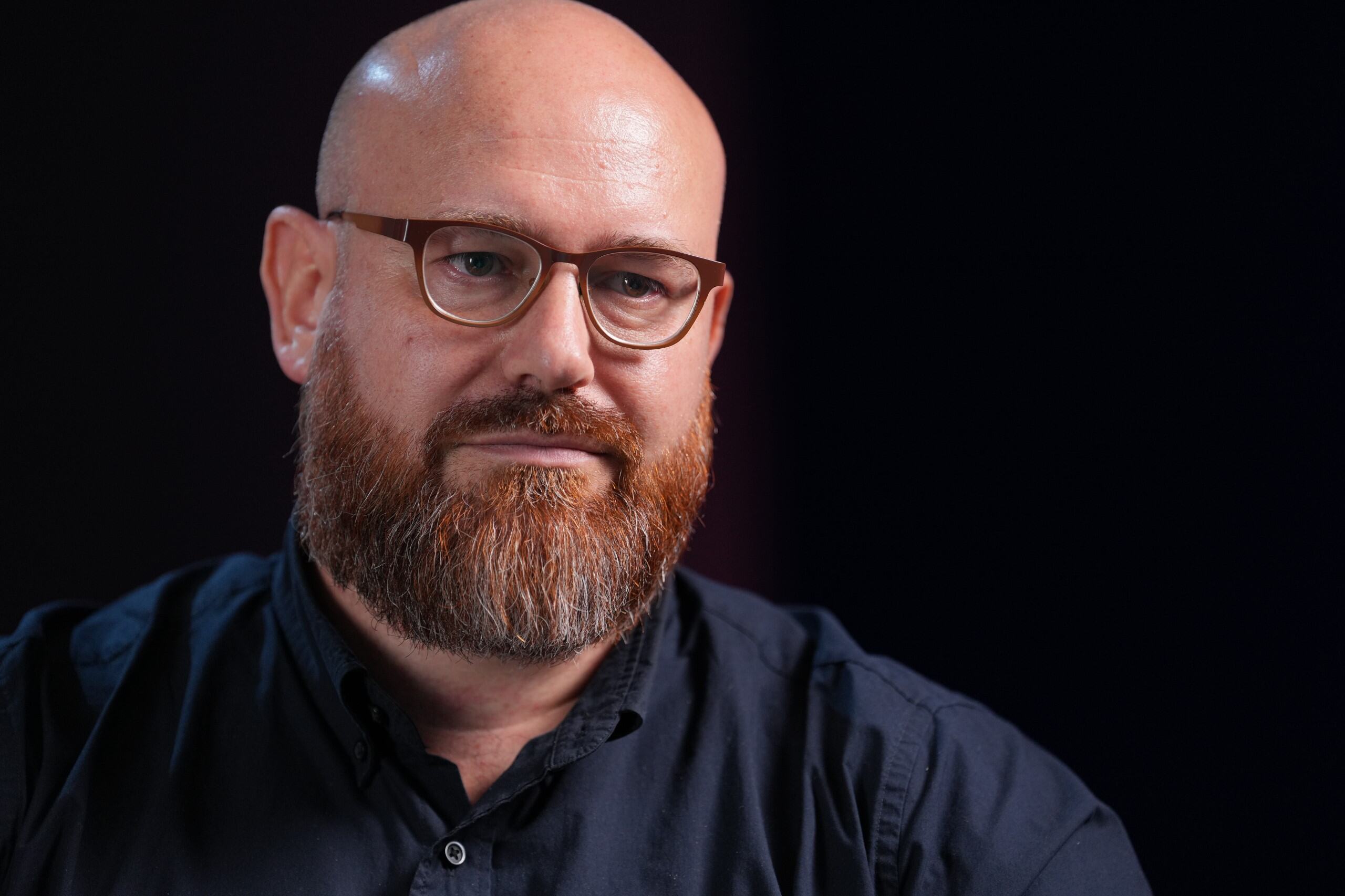 Homem careca com barba e óculos na frente de um fundo preto 