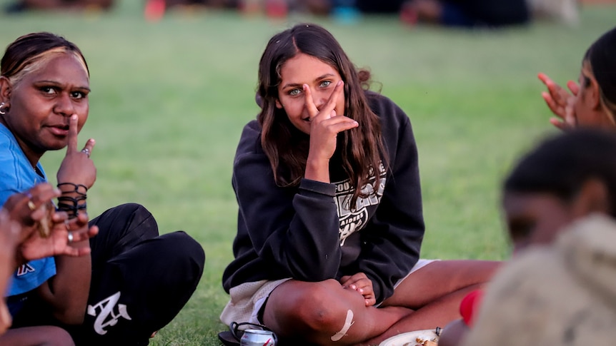 A young Aboriginal woman throws her fingers in front of her face, sitting on the grass among other young women