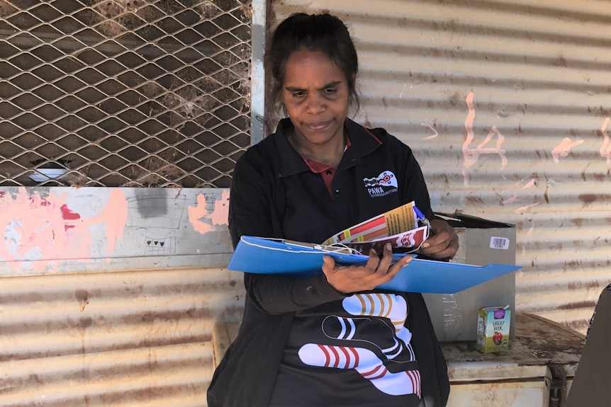 An young Aboriginal women with a ponytail dressed in a black polo shirt with a logo carries rolled posters comes out of a house