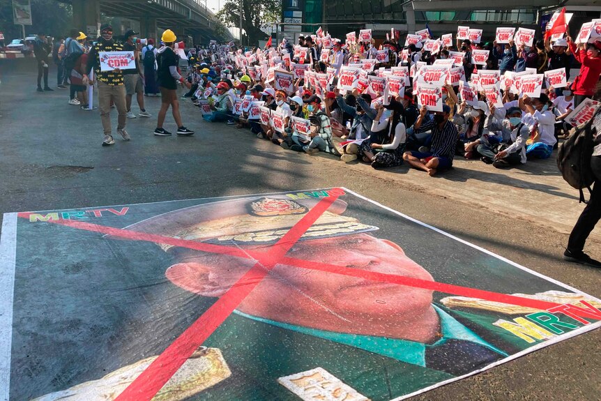 Hundreds of protesters sit near a large image that has an X mark on the face of a military commander.