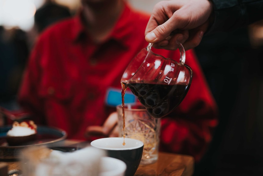 A close-up of a hand pouring a drink from a carafe into a cup. Diner wearing red shirt in background, out of focus.