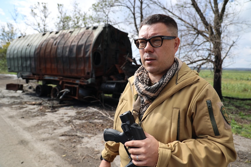 A man with short brown hair and thick black glasses stands in front of  ruined tank on a dirt road in Ukraine