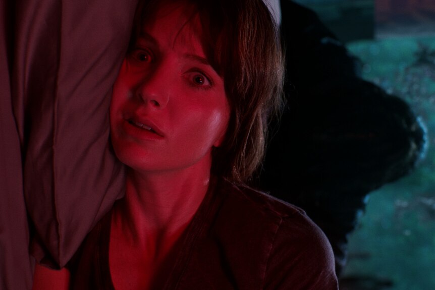 A woman with a fringe bathed in red light lies terrified on a bed, unidentifiable blue blob in the background