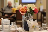 Flowers in a vase surrounded by glasses and a plastic drink bottle on a table at a nursing home, with a patient (unidentifiable)