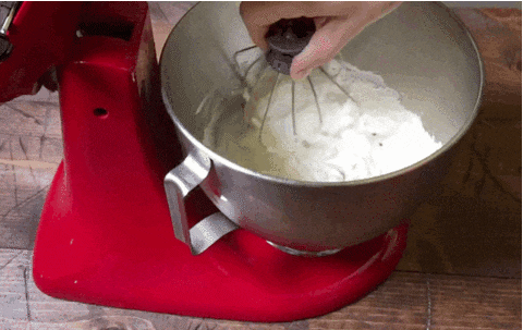 A hand lifts a wire whisk up showing how stiff the whipped cream is in the silver bowl.