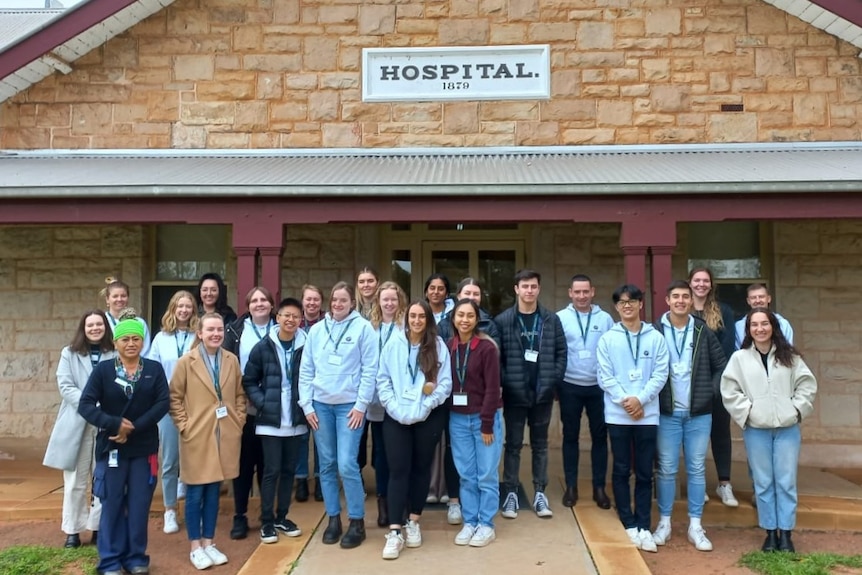 A group of students standing in front of a stone building, that has a sign that reads 'Hospital'