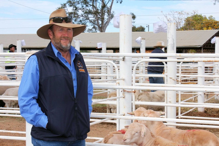 A farmer stands in front of a stockyard, grinning, with his hands in his pockets.