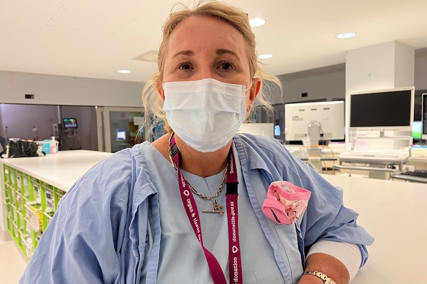 A woman wearing a mask and hospital scrubs looks at the camera. 