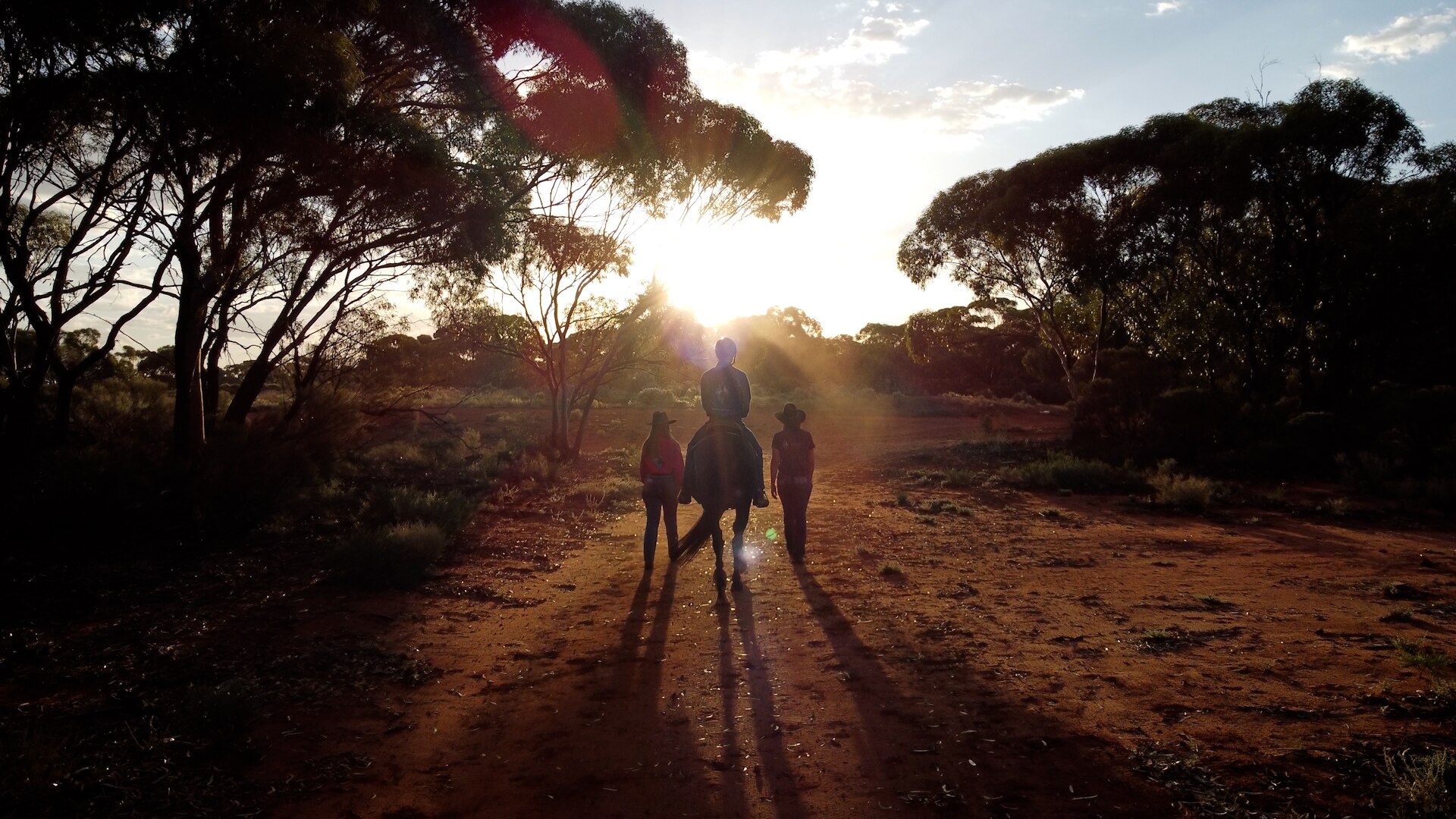 A teenager on a horse, two women either side, all walking into a golden sunset on a red dirt path.