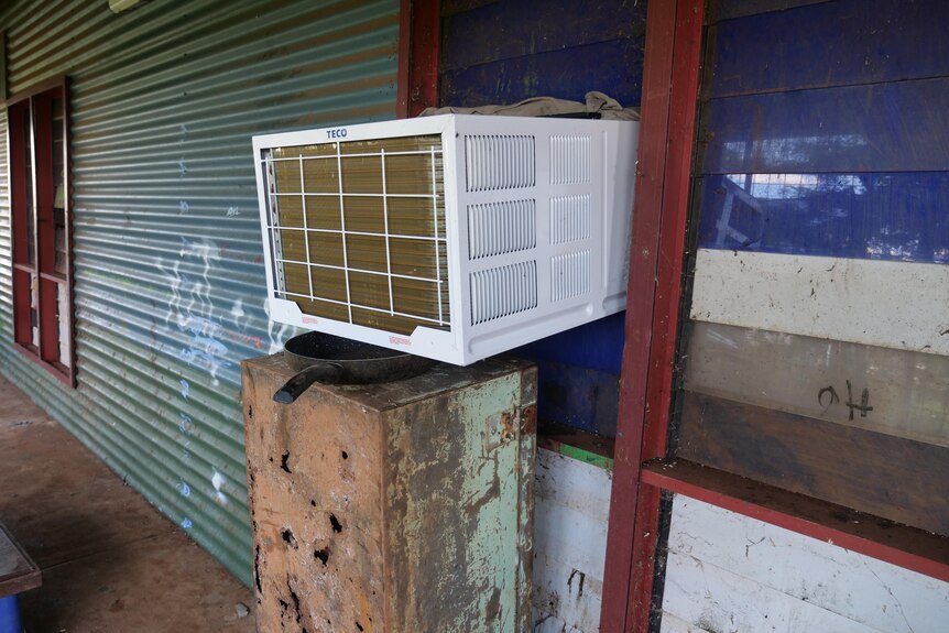 an air conditioner shoved through a window sitting on a frying pan supported by a metal cabinet