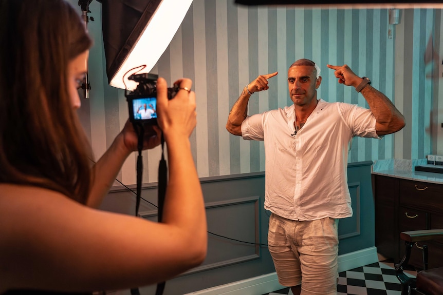 A man poses, flexing his biceps with both hands pointing to his head, while a woman takes his photograph on a digital camera