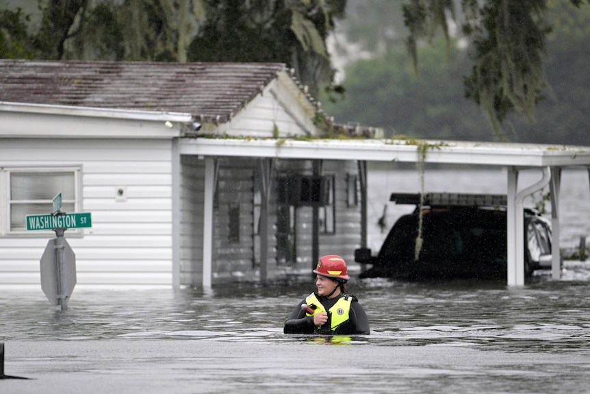 A woman wearing a bright helmet and hi-vis vest walks through floodwaters in front of wooden houses.