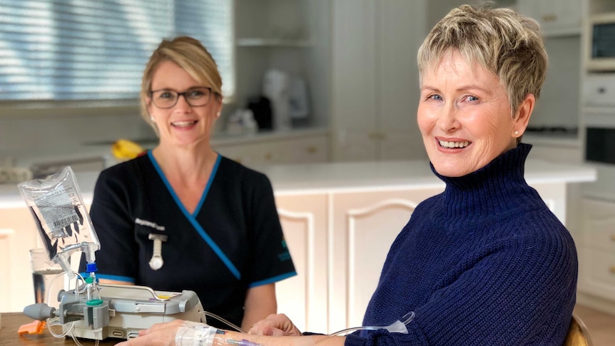 A woman sits at a dining table with an IV in her arm while a nurse looks on.