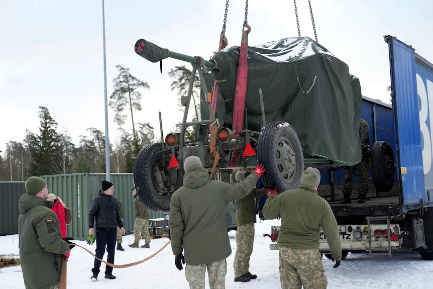 Men in camo uniforms guide a contraption hoisted by a crane to the snowy ground. 