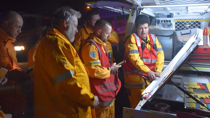 Queensland Rural Fire Brigade volunteers stand behind a vehicle at night assessing bushfire conditions.