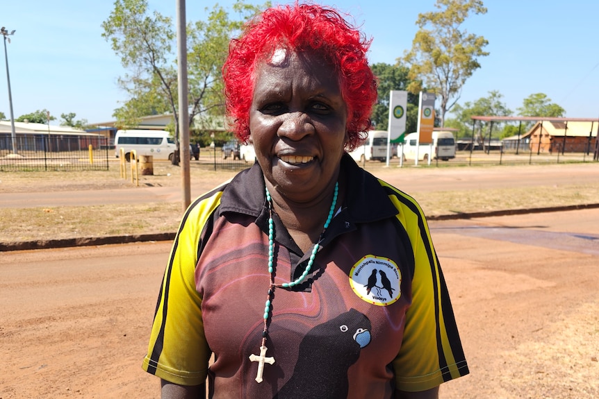 A woman smiles at the camera, the town of Wadeye is behind her