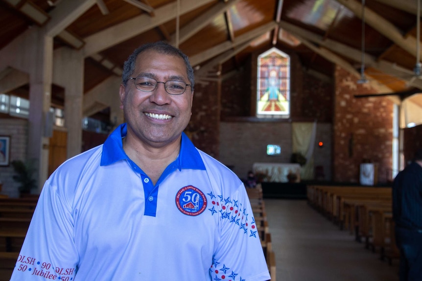 A man wearing glasses and a white and blue polo shirt smiles as he stands inside a church.