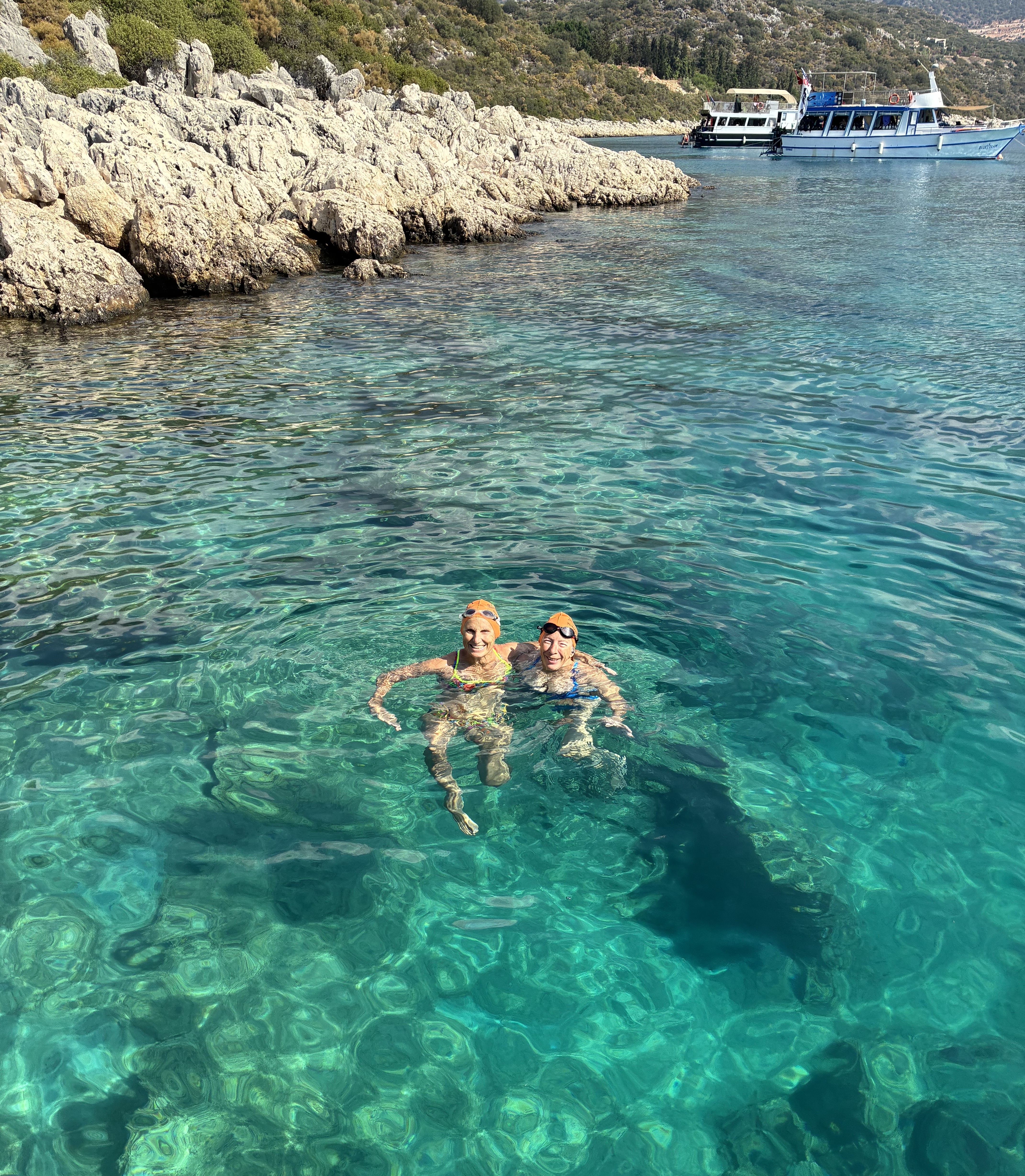 Duas mulheres em trajes de banho e toucas de natação flutuam em águas azuis claras com barcos e uma costa rochosa ao fundo.