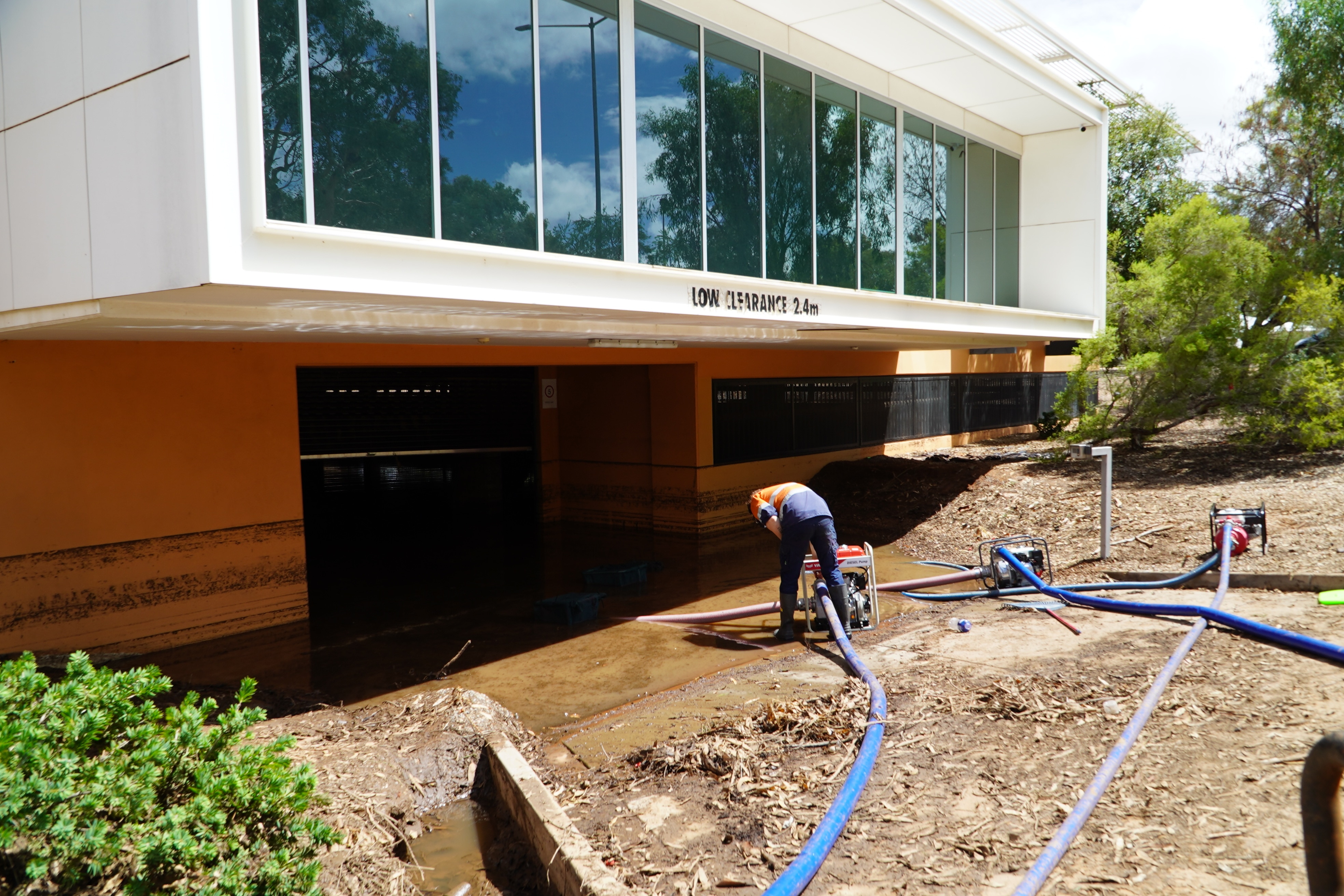 Um homem de calça azul e camisa laranja abaixa uma mangueira azul conectada a uma bomba na água que sai de um prédio laranja