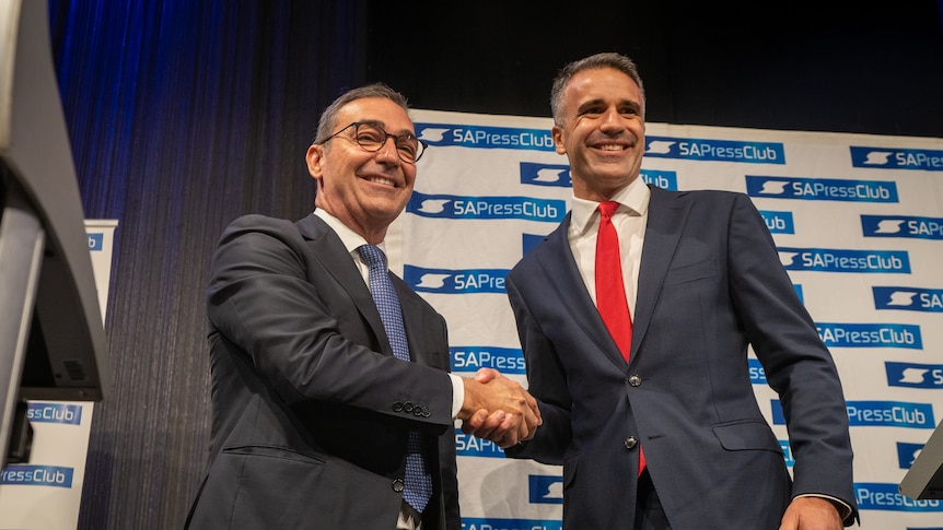 Two men in suits shake hands in front of a banner that reads SA Press Club