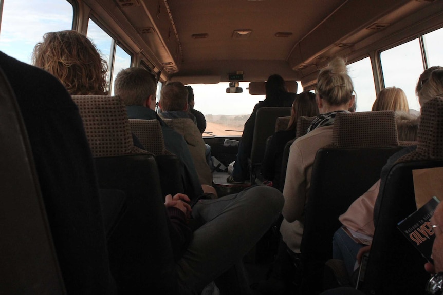 The bus is full with the back of teachers heads as they face a view of a dirt road through the front window
