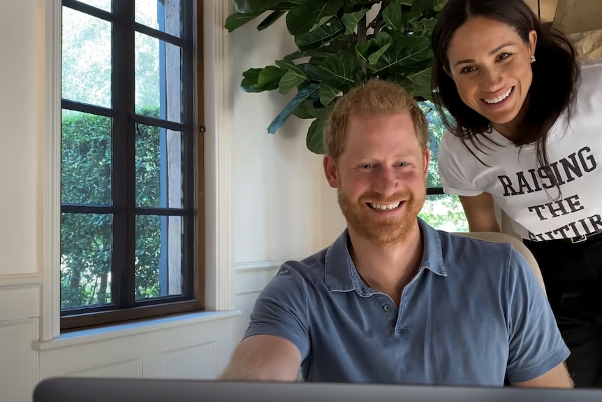 Prince Harry sits in a chair smiling at a screen. His wife Meghan is standing behind him also smiling next to a window