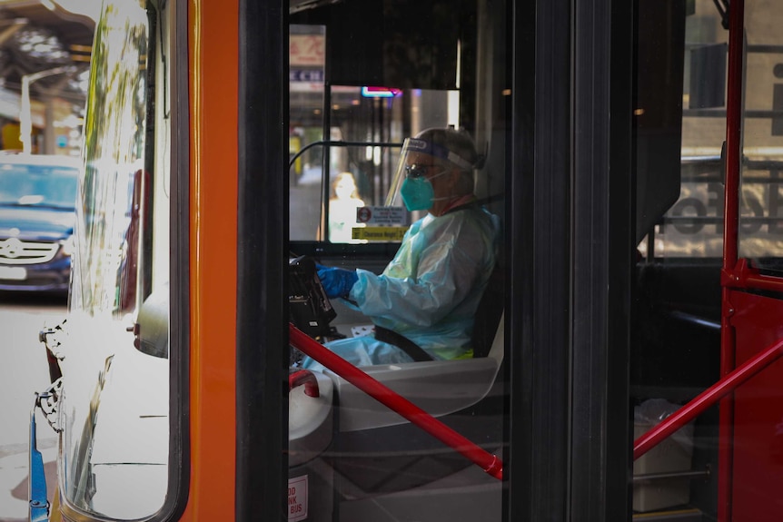 A bus driver sitting in the driver's seat wearing personal protective equipment including a mask, face shield and gloves