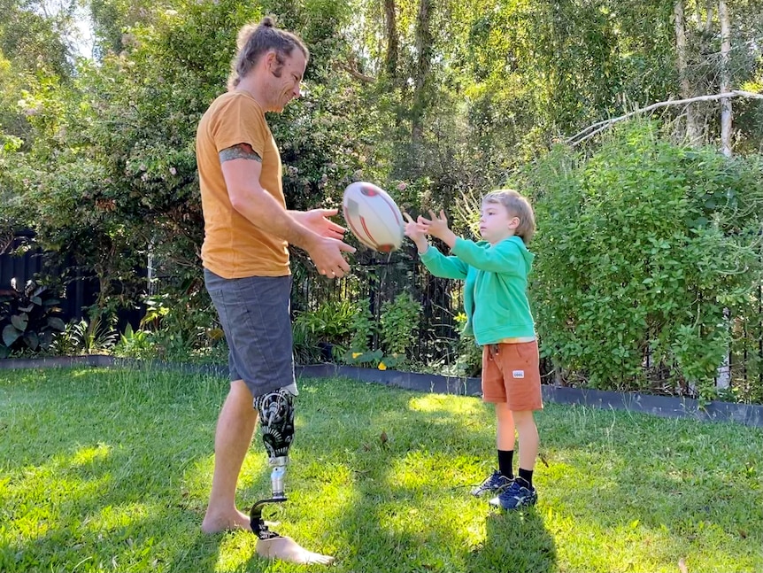Man with prosthetic leg throws footy with six-year-old son in the backyard